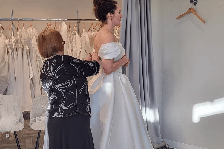 Bridal stylist adjusting the fit of a bride's off-the-shoulder satin wedding gown during an alterations appointment.
