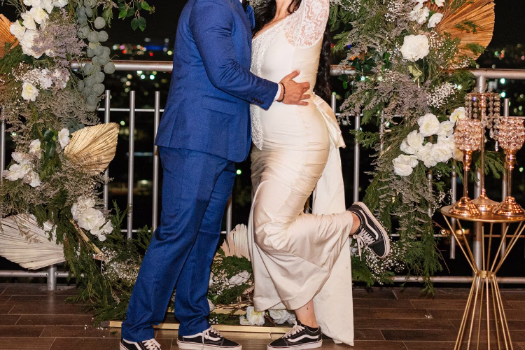 Newlywed couple posing in front of a floral wedding backdrop during their reception, showcasing a modern bridal look.