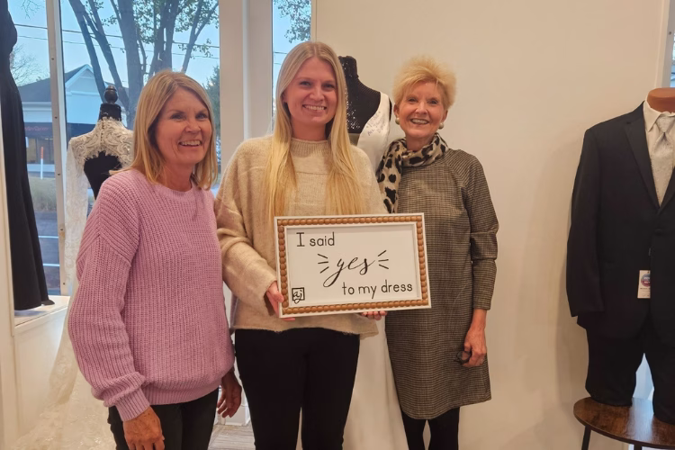 Bride-to-be holding a “I said yes to my dress” sign while posing with two family members inside a bridal boutique during a wedding dress shopping appointment.