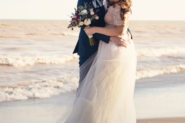 Bride and groom embracing on the beach, with the bride wearing a lightweight wedding dress ideal for a seaside wedding.