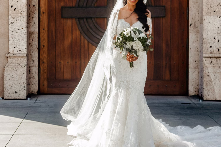 Bride wearing a lace fitted wedding dress with a long veil and bouquet, standing outside a church venue.