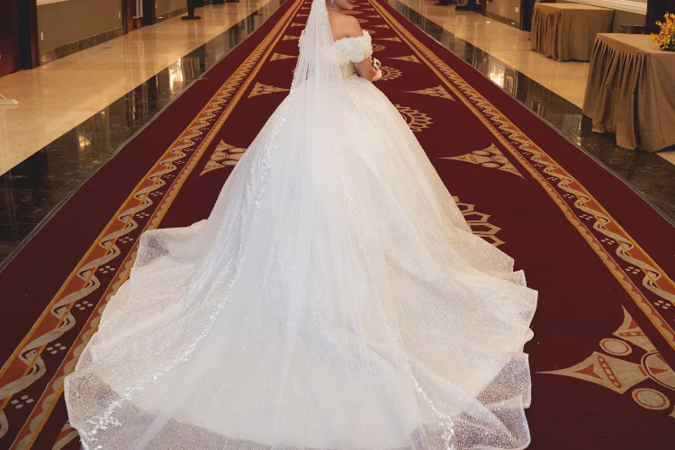Bride wearing a sparkling ball gown wedding dress with a long train and veil, standing in an elegant ballroom venue.