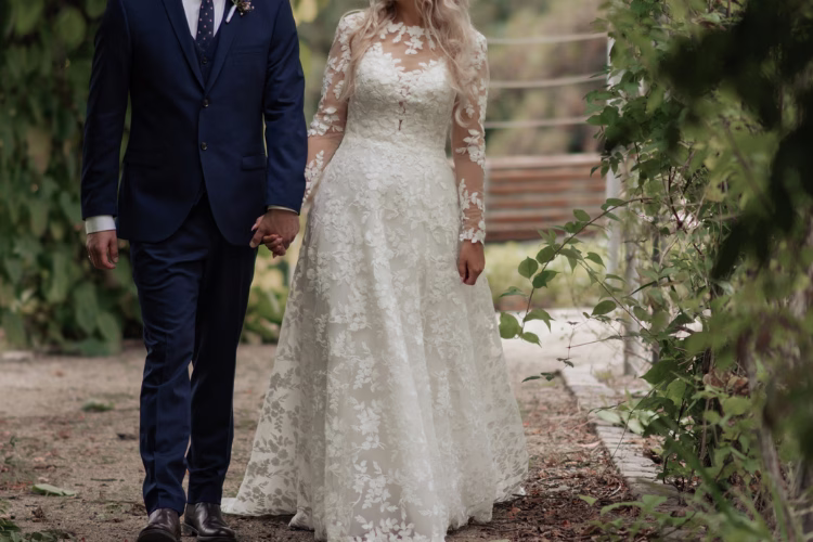 Bride and groom walking through a garden, with the bride wearing a floral lace wedding dress featuring long sleeves.