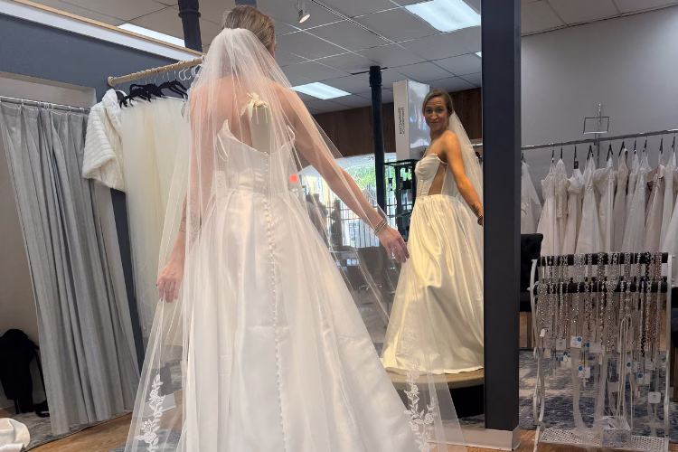 Bride admiring an A-line satin wedding dress with a long veil during her wedding dress shopping appointment.