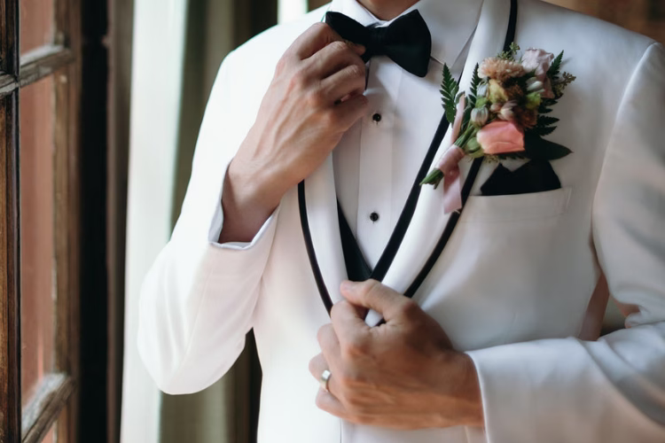 Close-up of a groom adjusting his black bow tie while wearing a white tuxedo jacket with black trim and a floral boutonniere.