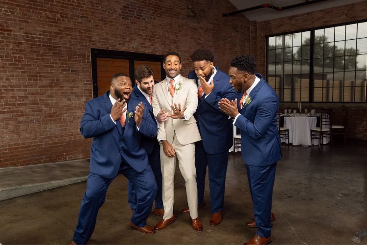 Groom in a cream suit surrounded by four groomsmen wearing navy blue suits in a brick-walled wedding venue.