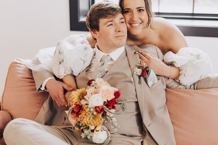 Groom in a beige suit sitting with his bride on a sofa, holding a colorful wedding bouquet.
