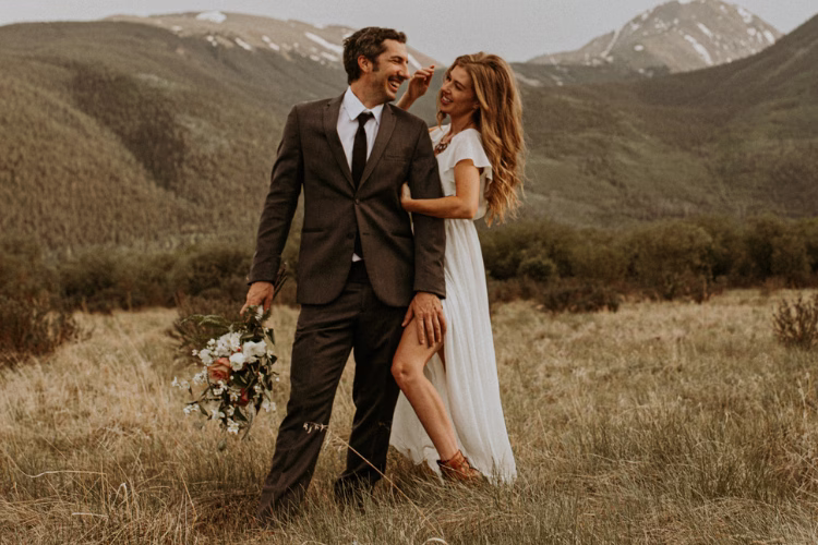Bride and groom standing in a mountain landscape, with the groom wearing a dark brown suit and the bride in a white dress.