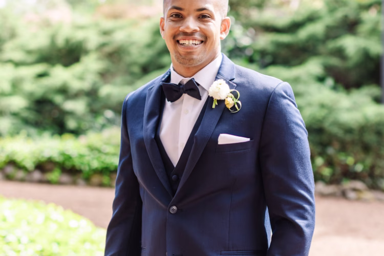 Smiling groom wearing a navy tuxedo with black lapels, black bow tie, white shirt, and floral boutonniere.
