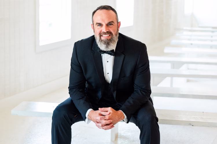 Groom with a beard sitting indoors in a black tuxedo, white dress shirt, and black bow tie.