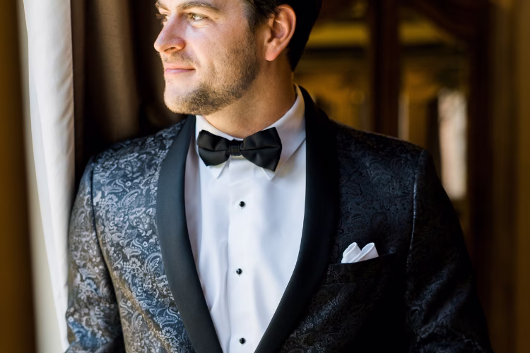 Groom wearing a black patterned tuxedo jacket with satin lapels, white dress shirt, and black bow tie.