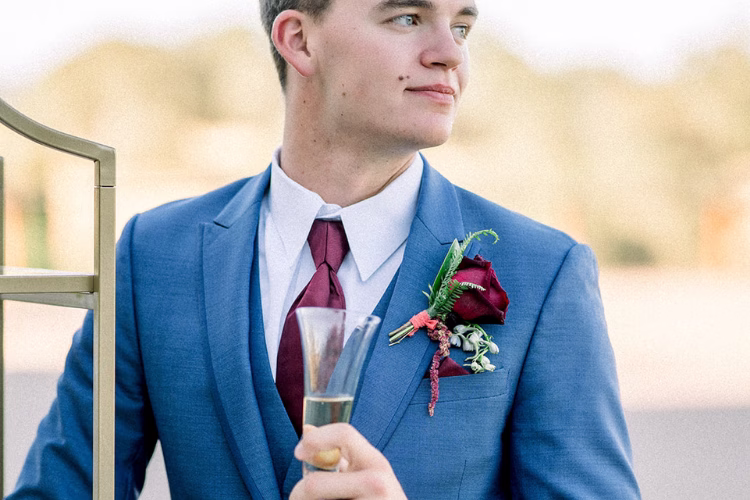 Groom wearing a bright blue suit with a burgundy tie and floral boutonniere while holding a champagne glass.