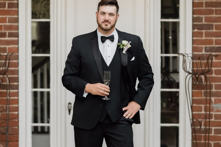 Groom wearing a black tuxedo with a matching vest, bow tie, boutonniere, and white pocket square while holding a champagne glass.