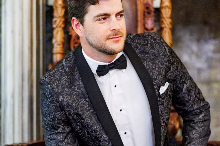 Groom wearing a patterned black tuxedo jacket with a black bow tie and white pocket square.
