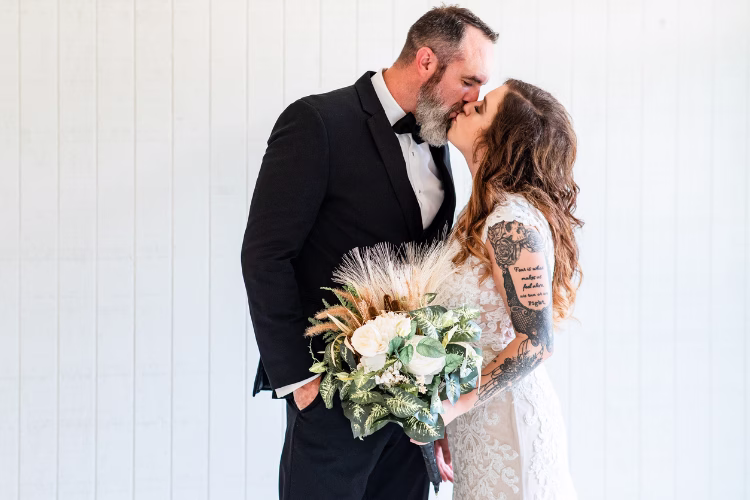 Groom in a black tuxedo kisses his bride while she holds a wedding bouquet.