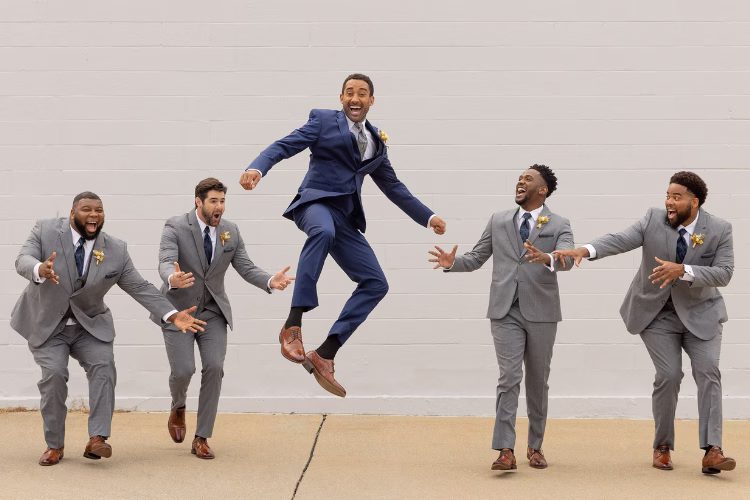 Groom in a blue suit jumps in the air while his groomsmen celebrate around him.