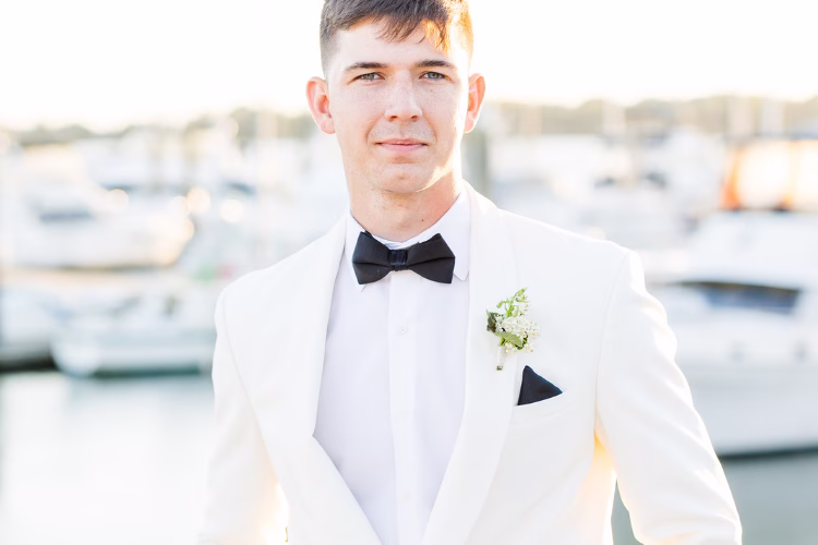 Groom wearing a white tuxedo jacket with a black bow tie and boutonniere at a waterfront wedding venue.