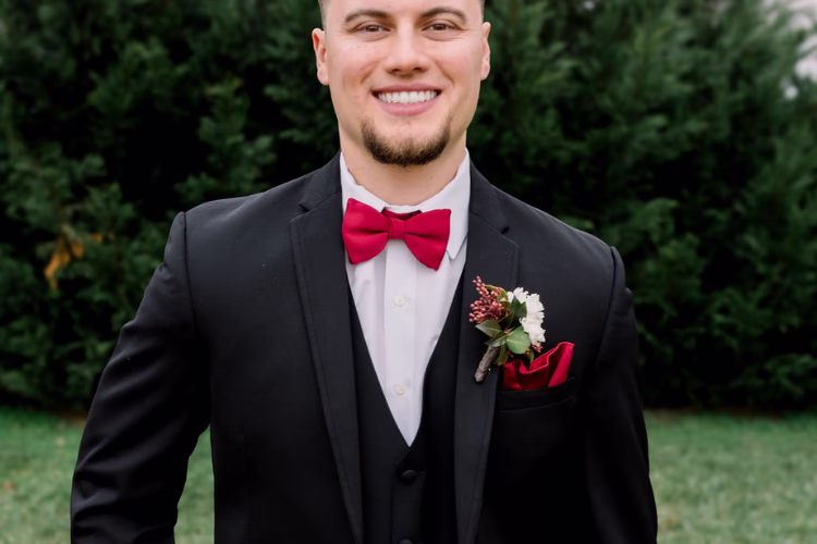Groom wears a black tuxedo with a red bow tie, pocket square, and boutonniere.
