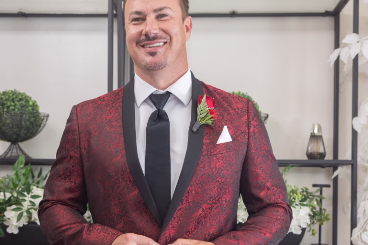 Groom in a black tuxedo and matching vest stands outside a brick home.
