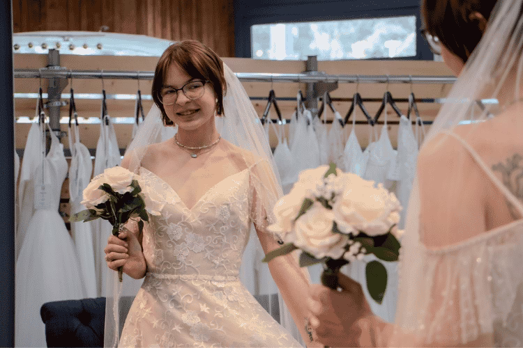Bride holding a bouquet while looking at her reflection in a wedding dress inside a bridal boutique