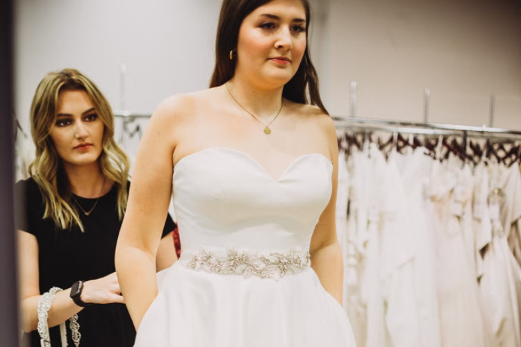 Bride wearing a strapless wedding gown while a bridal consultant adjusts the back of the dress