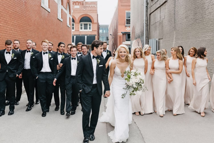 Wedding couple walking with bridesmaids in neutral dresses and groomsmen in black tuxedos.