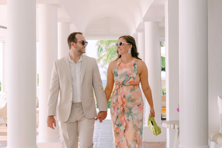 Couple walking hand in hand through a bright resort, with the man wearing a light beige suit and the woman in a floral dress.