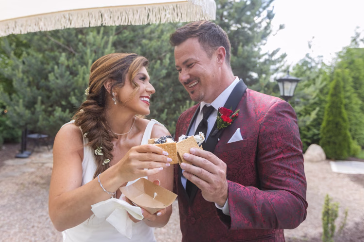 Bride and groom smiling as they share a dessert, with the groom wearing a burgundy patterned tuxedo jacket and boutonniere.